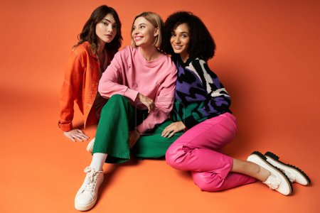 Three young, multicultural women in stylish clothes sit on the ground, posing for a fun picture against an orange backdrop.