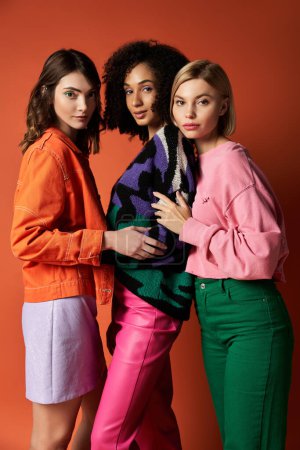 Three young, stylish women of different cultures standing together on an orange background, showcasing friendship and diversity.