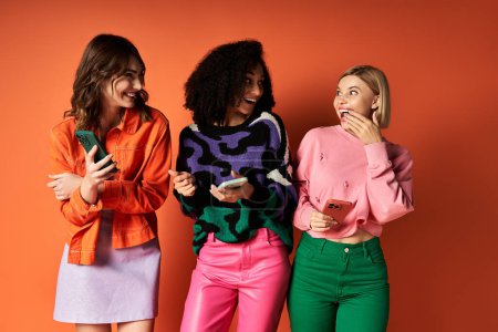 Three young women in vibrant outfits stand together on an orange background, showcasing diversity and friendship.