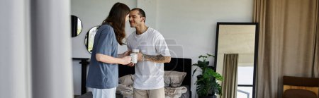 A young gay couple stands in a bedroom, holding mugs and looking lovingly into each others eyes.