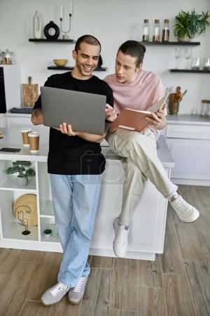 A young gay couple enjoys a cozy afternoon in their modern kitchen, one browsing on a laptop while the other reads a book.