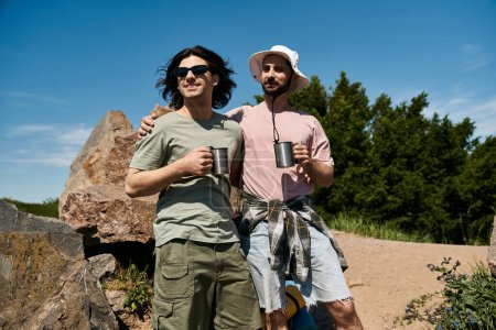 A gay couple enjoys a summer hike together, stopping for a moment to admire the view and share a drink.