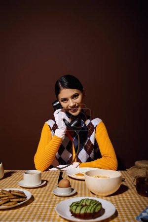Mujer con estilo en suéter amarillo en la mesa, hablando por teléfono rotatorio durante el desayuno.