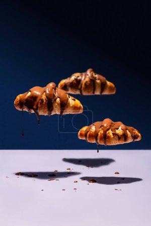 Foto de Nubes de croissant cubiertas de chocolate flotando y goteando sobre fondo azul - Imagen libre de derechos