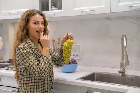 Téléchargez les photos : Joyeux jeune femme aux cheveux longs mangeant des raisins dans une cuisine - en image libre de droit