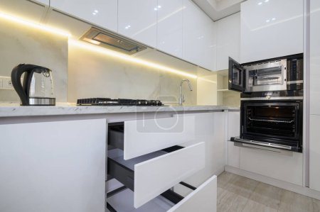 White modern kitchen with a stove, oven and microwave with open doors, glass electric kettle at worktop, drawers are retracted