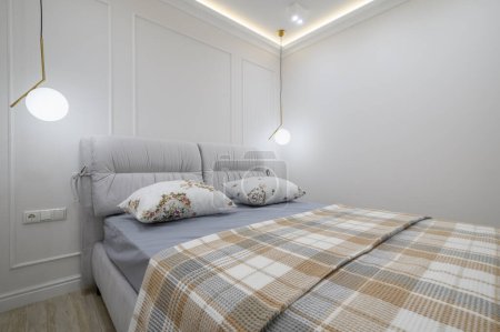 Photo for Closeup to double bed with a plaid blanket and pillows on it in a cozy bedroom with white walls and a light fixtures - Royalty Free Image