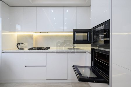 Foto de Cocina moderna blanca con fogones, horno y microondas con puertas abiertas, hervidor eléctrico de vidrio en la encimera. - Imagen libre de derechos