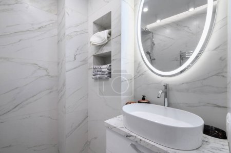Interior del baño blanco con azulejos de mármol en las paredes y un espejo redondo