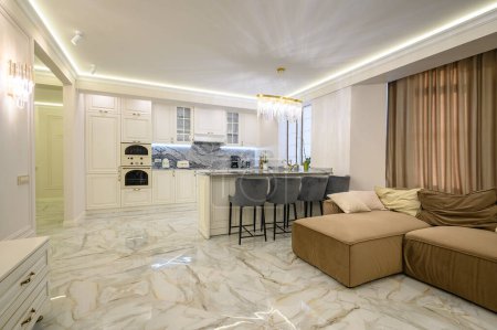 Foto de Open-concept studio apartment with classic kitchen in warm cream color and island for dining, featuring comfortable brown sofa - Imagen libre de derechos