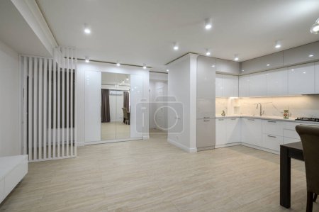 Photo for Large white room connected to hall and kitchen in a studio apartment, part of white kitchen furniture at right side - Royalty Free Image