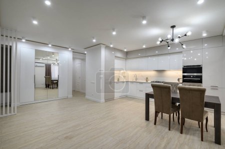 Photo for Interior of new spacious white kitchen after renovation in studio apartment, dining table and plenty of copy space, LED lighting in the sideboard - Royalty Free Image