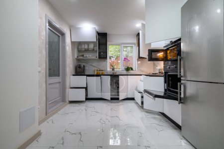 Téléchargez les photos : Une superbe cuisine avec un design blanc et moderne, un sol en marbre luxueux et des étagères coulissantes pour un accès facile. Les fours s'ouvrent. - en image libre de droit