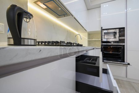 Téléchargez les photos : Cuisine moderne blanche avec cuisinière, four et micro-ondes avec portes ouvertes, bouilloire électrique en verre sur le plan de travail, tiroirs rétractés - en image libre de droit