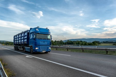 Téléchargez les photos : Big blue truck driving on the highway through forested landscape at sunset. - en image libre de droit