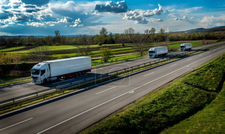 Highway transportation scene with three white transportation trucks in line on a rural highway under a beautiful blue sky