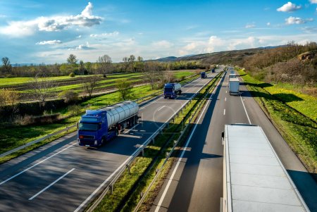 Schwere Highway-Transport-Szene mit Konvoi weißer Transport-LKWs in einer Richtung und blauen Tankschienen, die in der Gegenrichtung auf der ländlichen Autobahn unter einem schönen blauen Himmel passieren
