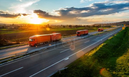 Téléchargez les photos : Trois camions de transport orange avec remorques sur une route asphaltée dans un paysage rural au coucher du soleil - en image libre de droit