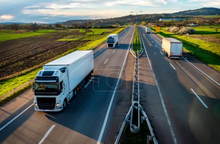 Foto de Escena de transporte pesado en una carretera con convoyes de camiones de transporte blanco que pasan por el camino opuesto en la carretera rural bajo un hermoso cielo azul - Imagen libre de derechos