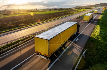Téléchargez les photos : Camions de transport avec remorques sur une route asphaltée dans un paysage rural à un beau coucher de soleil - en image libre de droit
