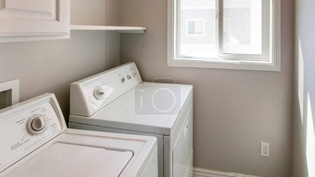 Panorama Laundry room interior with light gray wall and white cabinets and laundry units. There is a wall cabinet near the shelf on the left and single white rod near the sliding window.