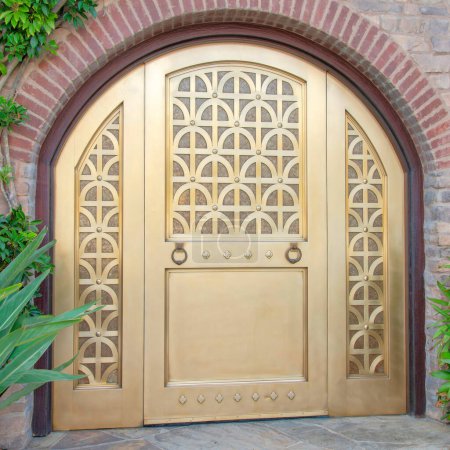 Photo for Square Arched gold door with two ring knockers at La Jolla, California. Entrance exterior with stone pavement and plants at the front of a door with arched bricks doorframe and stone sidings. - Royalty Free Image