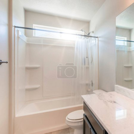 Téléchargez les photos : Square Sun flare Intérieur d'une salle de bain blanche avec des luminaires de salle de bain noirs et carrelage gris clair. Il y a une vanité avec dessus en marbre et miroir près des toilettes à côté de la baignoire de douche - en image libre de droit