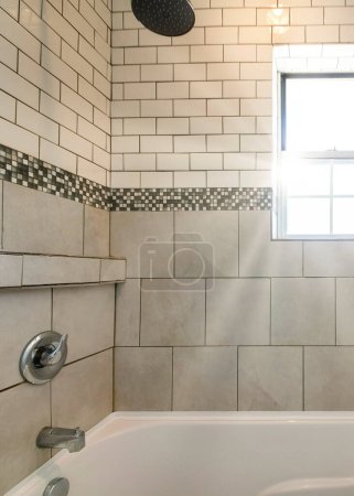 Photo for Vertical Sun flare Alcove bathtub shower combo with ceramic and subway tiles wall with mosaic tiles trim in the middle. Bathtub with wall mounted shower head and faucet near the single hung window at the front wall. - Royalty Free Image