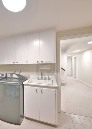 Vertikale Innenseite einer Waschküche mit offener weißer Tür und Blick auf den Flur. Waschmaschine und Trockner stehen neben dem Waschtisch unter den Schränken links neben den Schiebefenstern.