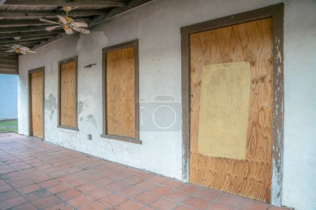 Foto de San Antonio, Texas- Exterior de una casa abandonada con puertas y ventanas cerradas. Porche de casa antigua con suelo de baldosas rojas bajo un techo de madera con ventiladores antiguos. - Imagen libre de derechos