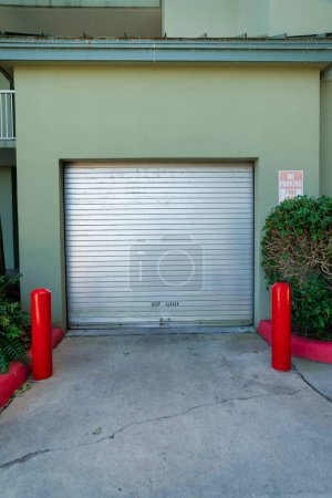 Foto de Destin, Florida puerta enrollable de acero de un garaje público en un hotel. Utilidad exterior garaje de un hotel con pared verde oliva pintado y una entrada de hormigón con dos postes rojos y plantas a la derecha. - Imagen libre de derechos