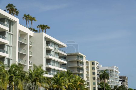 Foto de Fila de modernos edificios hoteleros en fila en Miami, Florida. Hay palmeras en la parte delantera de los edificios con árboles en las cubiertas de techo y barandillas de vidrio en los balcones contra el cielo. - Imagen libre de derechos