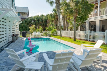 Destin, Florida- Small pool with flamingo pool float with legs in a yard with picket fence. There are four lounge arm chairs at the front facing the pool near the grass and palm trees on the right.
