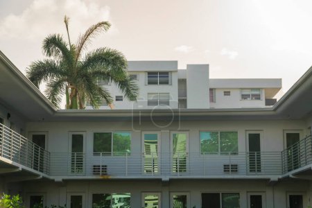 Foto de Fachada de un apartamento con puertas y ventanas reflectantes en Miami, Florida. Edificio de apartamentos exterior con terraza en la parte delantera de las puertas y vistas al árbol y edificio más alto en el fondo. - Imagen libre de derechos