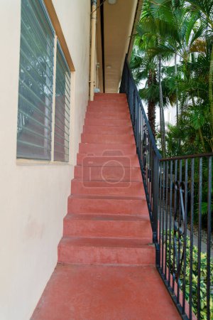 Photo for Stairs with red concrete steps and steel railings beside the building wall at Miami, Florida. Entrance at the side of the building with jalousie windows on the left and yard with trees on the right. - Royalty Free Image