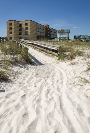 Foto de Camino en la arena que se dirige a un paseo marítimo en medio de dunas de arena en la parte delantera del hotel- Destin, FL. Camino de madera entre las hierbas en las dunas de arena blanca y vista de un hotel al fondo. - Imagen libre de derechos