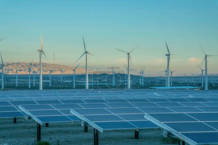Desert wind farm near mountains with solar panels and wind turbines in California. View of solar panels and windmills during sunset and a background of mountain range sky at the back.