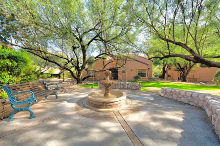 Téléchargez les photos : Petit parc avec banc et fontaine d'eau dans un lotissement à Tucson, Arizona. Il y a un mur de pierre sur la droite et une pelouse avec des arbres près de la passerelle menant aux maisons italiennes à l'arrière. - en image libre de droit