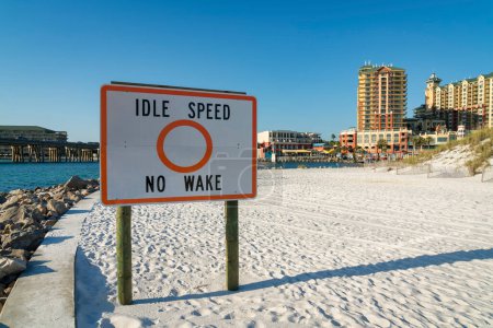 Téléchargez les photos : Idle Speed No Wake signe sur un sable blanc au bord de la plage près des hôtels à Destin, Floride. Panneau près de l'eau et le pont sur la gauche et les bâtiments de l'hôtel sur la droite. - en image libre de droit