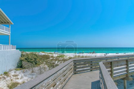 View of the beach with tourists on the shore from a boardwalk at Destin, Florida. There is a view of a beach house on the left with balconies near the tall grasses at the side and a waterfront view.