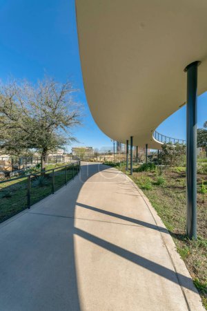 Foto de Austin, Texas- Paseo en medio de pastizales verdes bajo un puente en Waterloo Park. Sendero de hormigón con valla de alambre de metal en el lado y una vista de los árboles y el cielo en el fondo. - Imagen libre de derechos
