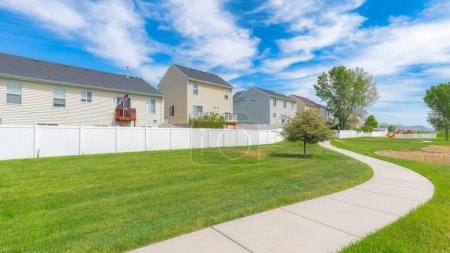 Photo for Panorama Whispy white clouds Concrete pathway in the middle of a green field with trees in a residential area at Utah valley. There are houses with decks and wood sidings at the back of white vinyl fence. - Royalty Free Image