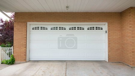 Panorama Porte de garage sectionnelle blanche avec panneaux de verre en haut. Extérieur d'une maison avec revêtement en briques brunes et une allée en béton à l'avant du garage.