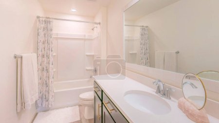 Panorama Intérieur de salle de bain blanche avec miroir rond sur une pierre au comptoir d'un lavabo élégant. Il y a une baignoire douche avec panneau mural en acrylique et rideau près des toilettes avec tapis blanc sur le sol en bois.