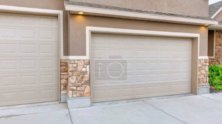 Panorama Garage extérieur à trois voitures avec revêtement brun clair. Il y a deux portes de garage sectionnelles avec allée en béton et un revêtement en béton et placage de pierre.