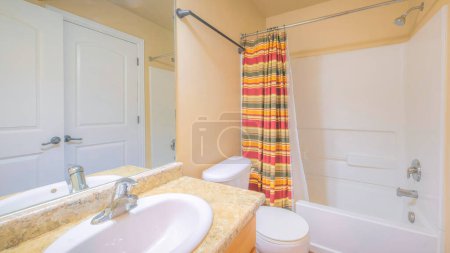 Téléchargez les photos : Panorama Intérieur de salle de bain avec deux portes blanches et des murs de couleur chaude. Il y a un lavabo avec comptoir en granit et miroir près des toilettes à côté de la douche de baignoire avec pomme de douche murale. - en image libre de droit