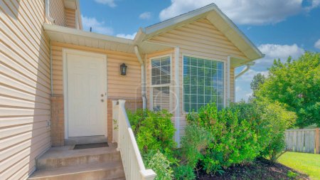 Photo for Panorama White puffy clouds White front door near the vinyl wood siding of a house in Utah. House exterior with shrubs near the bay windows beside the railing of the doorsteps with wall lamp and a view of a lawn with fence. - Royalty Free Image