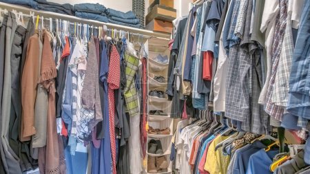 Photo for Panorama Walk-in closet with hanging linen shoe rack and clothes. There is a wall mounted shelves with shoe boxes and denim pants on top and a hanging clothes on the rods and a plastic drawers below. - Royalty Free Image