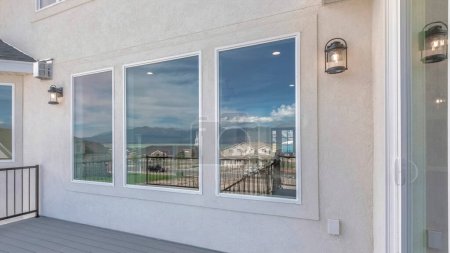 Panorama Deck of a house with sliding glass windows and open wall lamps. Exterior of a house with glass picture windows and a deck with wood planks flooring and metal railing.