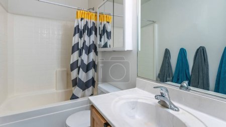 Téléchargez les photos : Panorama Intérieur d'une petite salle de bain avec des serviettes grises et bleues suspendues au mur. Il y a un lavabo avec miroir près de la toilette avec armoire miroir à côté de la baignoire de douche avec panneau mural motif carrelage. - en image libre de droit